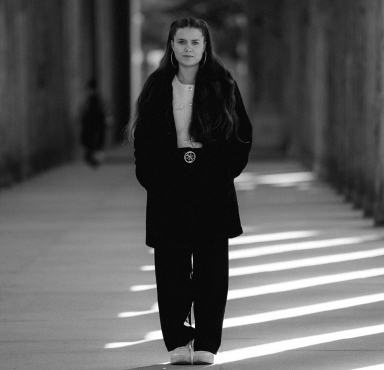 Foto: Junge Frau in schwarzem Outfit steht allein in einem lichtdurchfluteten Gang.